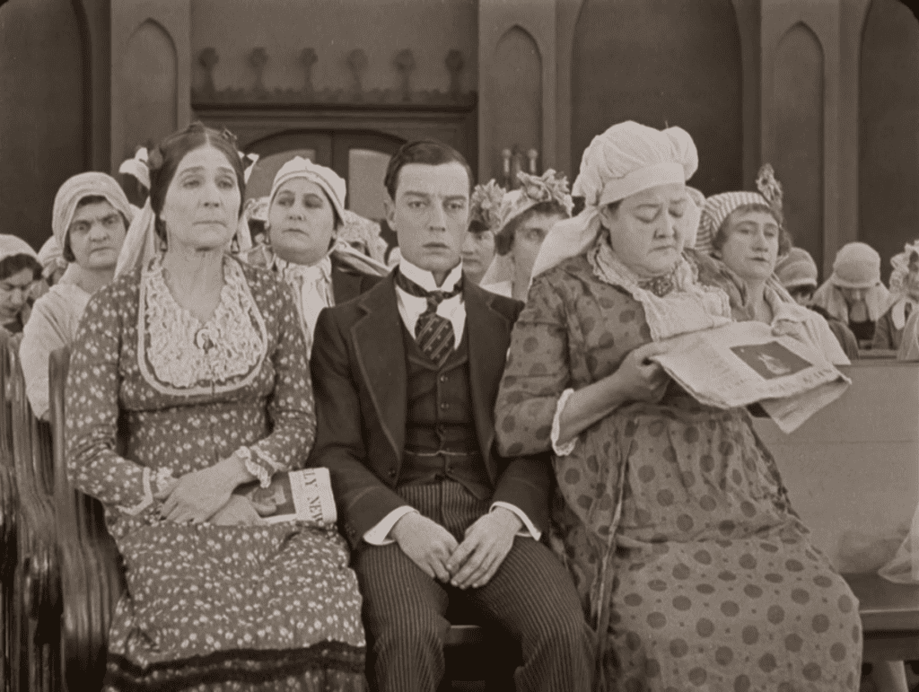 Buster Keaton sitting in a church with a crowd of women hoping to marry him, from the film Seven Chances.