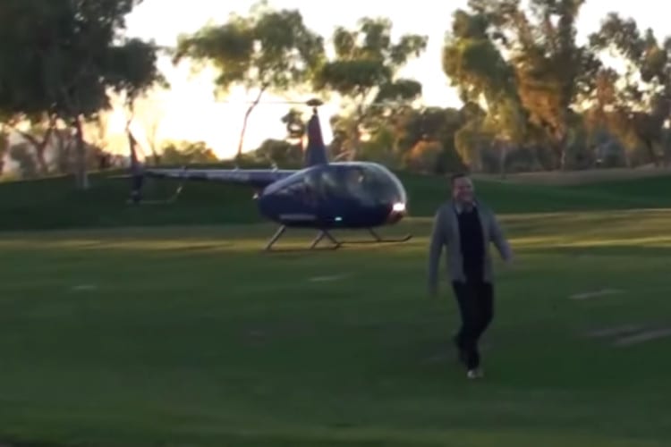 Man walks across a golf course green, a helicopter behind him.